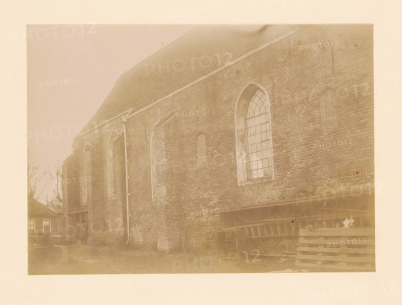 North side of the St. Lambertus Church in Wilsum, photograph, annotation: 'Wilsum', annotation: 'Wilsum North side of the Dutch Reformed Church', Vollenhove, 1897, photographer, Attributed to, paper, pasteboard, albumen print, height 164 mm x width 228 mm, church (exterior), Wilsum, St. Lambertus Church (Wilsum), A faded sepia photograph showing the side of an old brick building with a steep tiled roof and several tall arched window openings, one of which contains a multi-pane window, and others appearing filled or bricked in; a vertical downspout runs along the wall; the brickwork shows weathering and patches of lighter mortar; at the ground level there are stacked wooden pallets and a small open doorway, and in the left background there are bare tree branches and the corner of a smaller building with visible windows.