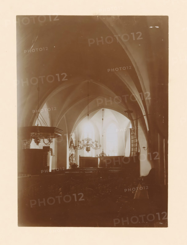Interior of the St. Nicholas Church in Wijhe, photograph, annotation: 'Wijhe', annotation: 'Dutch Reformed Church in Wijhe Aug 1911', St. Nicholas Church (Wijhe), 1911, photographer, Wijhe, Attributed to, paper, pasteboard, daylight collodion silver print, height 213 mm x width 150 mm, interior of church, A sepia-toned interior scene showing a vaulted ceiling with pointed arches and ribbing, hanging light fixtures and chandeliers suspended from long rods, tall arched windows at the far end allowing light to filter through, dark wooden furnishings including a raised pulpit with ornate detailing on the left and pews extending toward the foreground, decorative wall-mounted lamps on the right, and sculptural or carved elements near the front beneath the windows.