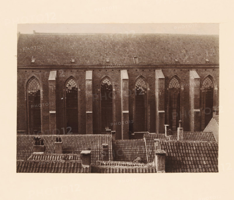 South side of the Broerenkerk in Zwolle, photograph, annotation: 'Broerenkerk, Zwolle', annotation: 'Broerenkerk in Zwolle, South side, June 1909', Zwolle, 1909, photographer, Attributed to, paper, pasteboard, albumen print, height 153 mm x width 208 mm, church (exterior), A sepia-toned photograph showing the upper facade of a large brick building with tall, narrow arched windows set between vertical brick pilasters, a tiled pitched roof above with small rectangular openings, and in the foreground a series of overlapping clay-tiled rooftops with chimneys and vents visible.