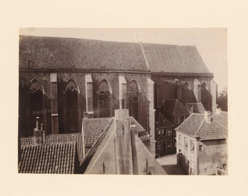 South side of the Broerenkerk in Zwolle, photograph, annotation: 'Broerenkerk in Zwolle, South side, June 1909', annotation: 'Broerenkerk, Zwolle', Zwolle, 1909, photographer, Attributed to, paper, pasteboard, albumen print, height 151 mm x width 218 mm, church (exterior), A sepia-toned photograph showing the upper portions of several buildings clustered together, dominated by a large brick structure with tall arched windows and a steep tiled roof, nearby smaller buildings with steep, tiled roofs and chimneys, visible rooflines and ridges creating angled shapes, textured roof tiles and brick walls, vertical downpipes and buttresses on the large building, shadowed window openings and panes in some of the facades, and a pale blank border surrounding the image.