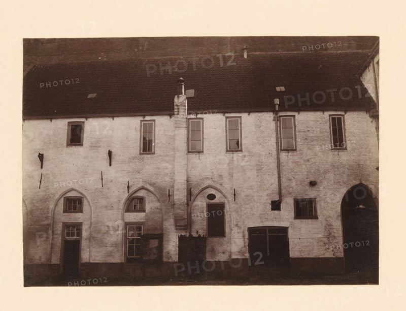 Monastery near the Broerenkerk in Zwolle, photograph, annotation: 'Broerenkerk in Zwolle, cloister north of the church in the barracks June 1909', annotation: 'Broerenkerk, Zwolle', Zwolle, 1909, photographer, Attributed to, paper, pasteboard, albumen print, height 155 mm x width 221 mm, cloisters, monastery, Broerenkerk, A faded, sepia-toned photograph of a brick building facade with a steep tiled roof, small roof vents, and a central chimney running down the wall; the upper wall has tall, narrow windows with muntins, while the lower wall features arched recesses, rectangular windows, and doorways of varying sizes; vertical downspouts and metal fixtures are attached to the exterior, and there is a dark arched passage visible at one side.