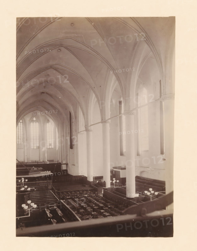 Interior of the Broerenkerk in Zwolle, photograph, annotation: 'Broerenkerk in Zwolle, Interior June 1909, viewed towards the choir', annotation: 'Broerenkerk, Zwolle', Broerenkerk, 1909, photographer, Zwolle, Attributed to, paper, pasteboard, albumen print, height 220 mm x width 157 mm, interior of church, A high vaulted interior with pointed arches and ribbed ceilings supported by tall smooth columns, large arched windows along the side letting in diffuse light, rows of wooden pews and chairs arranged in a central floor area, low wooden partitions creating aisles and enclosures, several multi-globe light fixtures mounted on stands, a raised area at the far end with larger windows behind it, and ornamental capitals where arches meet the columns.