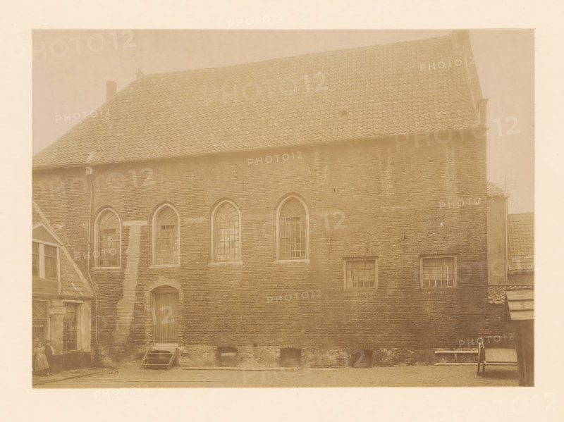 View of the synagogue in Zwolle, photograph, annotation: 'Synagoge, Zwolle', annotation: 'Zwolle synagogue March 1899 (O.IJ. is being demolished', Zwolle, 1899, photographer, Attributed to, paper, pasteboard, albumen print, height 166 mm x width 233 mm, the Synagogue (as building and place of worship, Jewish religion), A sepia-toned photograph of a large brick building with a steep tiled roof and several arched windows set into the upper wall, rectangular windows lower down, and a tall wooden door with a small stepped platform in front; a narrow vertical downpipe runs along the facade, and low arched openings sit at the base of the wall; to the left a smaller gabled structure with visible windows and a woman standing near its entrance, and to the right a partial view of another tiled roof and a wooden bench or cart against the wall.