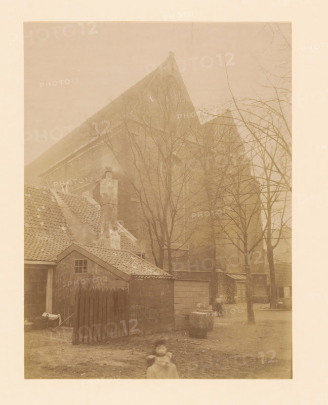 View of the Broerenkerk in Zwolle, photograph, annotation: 'Zwolle', annotation: 'Boerenkerk te Zwolle (O.IJ.)', Zwolle, 1899, photographer, Attributed to, paper, pasteboard, albumen print, albumendruk, height 228 mm x width 170 mm, church (exterior), Broerenkerk, A faded sepia photograph showing rooftops and a large steep gabled building partially obscured by leafless trees, with tiled roofs and a chimney in the foreground, a small wooden shed with a single window and a fenced gate, a dirt yard with scattered objects including a barrel and a low stone, a person standing near the trees in the middle distance, and a blurred child in the foreground.