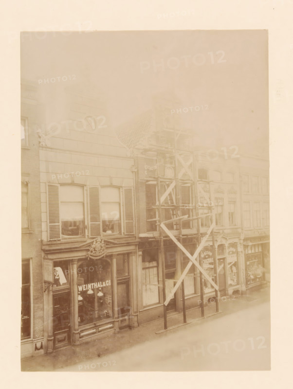 Diezerstraat 43 in Zwolle, photograph, annotation: 'Zwolle', annotation: 'Woonuis Diezerstraat 43 Zwolle (O.IJ.) Dec: 1905', Zwolle, 1905, photographer, Attributed to, paper, pasteboard, albumen print, height 230 mm x width 168 mm, urban housing, street, A faded sepia photograph of a row of multi-story storefront buildings with large windows and decorative architectural details, one facade showing shuttered upper windows and a triangular pediment with an emblem, another facade partially covered by wooden scaffolding with diagonal supports and vertical posts, carved ornamental moldings above the ground-level shop windows, display windows revealing hanging lamps and interior shelving, narrow sidewalks in front of the buildings, and a pale sky above.