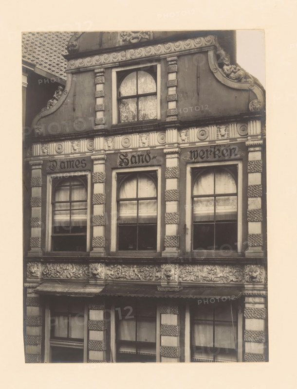 Façade of Sassenstraat 33 in Zwolle, photograph, annotation: 'Zwolle', annotation: 'Zwolle. House Sassenstraat 33. detail. Oct 1907', Zwolle, 1907, photographer, Attributed to, paper, pasteboard, gelatin silver print, developed gelatin silver print, height 220 mm x width 161 mm, urban housing, façade (of house or building), A sepia-toned photograph of a building facade showing ornate architectural details with three tall arched windows framed by decorative pilasters, patterned stonework on the pillars, carved floral and foliate friezes between the floors, and sculpted faces and reliefs integrated into the ornamentation; the upper section includes a rounded window with patterned curtains and a curved pediment flanked by reclining sculpted figures, while the lower sections show window panes with blinds or curtains and a projecting awning above the ground-floor opening.