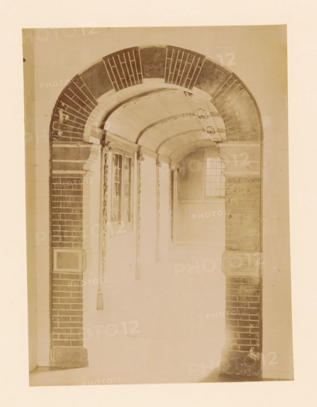 Covered hall in the Women's House in Zwolle, photograph, annotation: 'Zwolle hallway in the women's house on the Melkmarkt Sept 1893', annotation: 'Zwolle', Zwolle, 1893, photographer, Attributed to, paper, pasteboard, albumen print, height 232 mm x width 171 mm, vestibule, hall, old-people's home, A sepia-toned photograph of an arched corridor framed by brick and stone columns, showing a curved vaulted ceiling with decorative moldings and carved rosettes, a section of a window with small panes at the far end, ornate pilasters and carved vertical embellishments along the walls, and a floor and lower walls that fade toward the foreground.