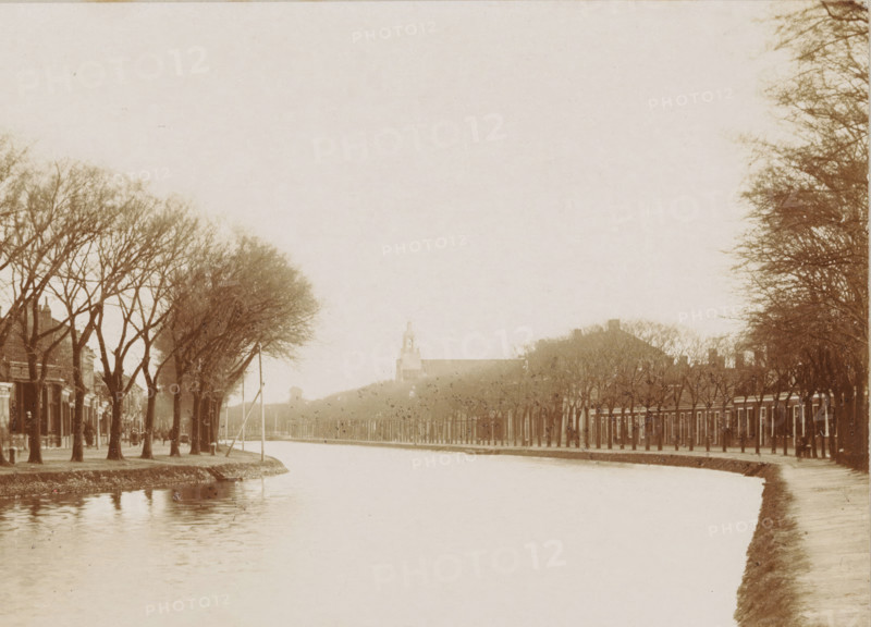 Canal in Den Helder, cabinet photograph, collector's mark: 'Vosmaer', number: '0.50', photographer: Anna Clasina Leijer, Den Helder, 1890 - 1910, Anna Clasina Leijer, photographer, paper, pasteboard, baryta paper, daylight collodion silver print, height 105 mm x width 163 mm, canals, waters (in city), A wide canal runs through the center of the scene with calm water reflecting pale tones, bordered on both sides by rows of bare deciduous trees lining paved paths and streets. On the left bank there are closely spaced houses and buildings partly obscured by tree trunks, and a narrow strip of grass at the water's edge. On the right bank a continuous line of uniform buildings extends into the distance beneath another avenue of trees, and a raised embankment slopes down to the canal. In the background a larger building with a tower rises above the treeline, appearing softened by distance and pale light. The overall image has a muted, sepia-toned palette and a soft, hazy atmosphere.