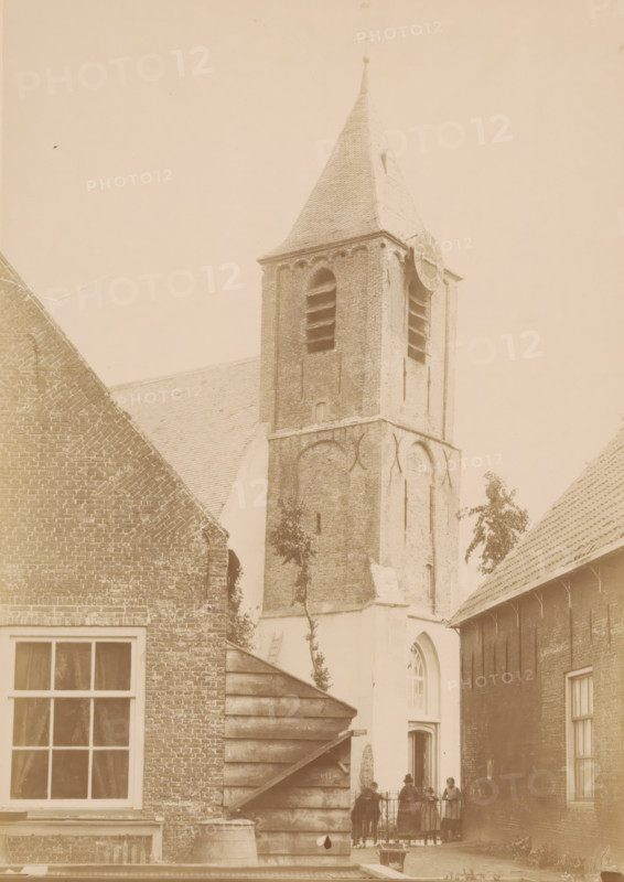 Tower of the Village Church of Nootdorp, photograph, annotation: 'Tower of the church of the Dutch Reformed congregation in Nootdorp, June 1891', annotation: 'Dutch Reformed Church, Tower, June 1891, Nootdorp', Nootdorp, 1891, photographer, Attributed to, paper, pasteboard, albumen print, height 239 mm x width 169 mm, parts of church exterior and annexes: tower, street (city(-scape) with figures, staffage), A tall brick church tower with a pointed roof and a circular clock face on one side rises between adjoining brick buildings, showing arched openings and louvers on the upper section and decorative brickwork on the lower section; in front of the tower a small courtyard area is visible with a wooden stepped structure and a barrel, a window with panes to the left, and a group of people standing near a gate by an arched doorway beneath the tower, while tiled roofs and a few small trees frame the scene.
