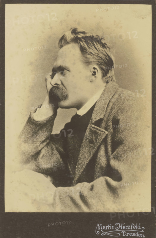 Portrait of Friedrich Nietzsche, photograph, annotation: 'Nietzsche', photographer: Martin Herzfeld, Dresden, c. 1882, 1910, Saxony, paper, pasteboard, daylight gelatin silver print, height 143 mm x width 101 mm, height 168 mm x width 109 mm, Person, Friedrich Nietzsche, historical persons, A sepia-toned portrait of a man shown in profile looking to the left, with a high forehead and swept-back hair, a thick mustache, and a hand resting against his cheek; he wears a collared shirt, a dark tie or vest beneath, and a textured jacket with a visible lapel and pocket, all set against a plain, lightly speckled background with a dark border around the image.