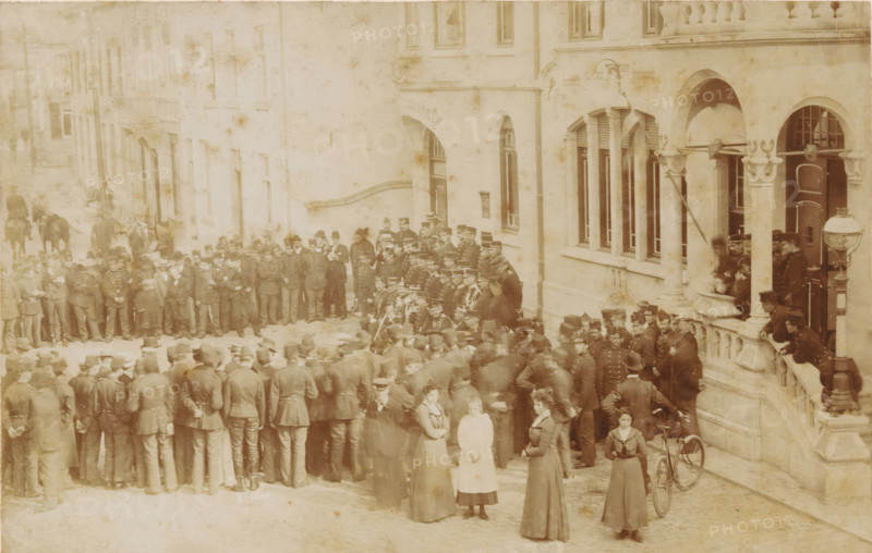 Soldiers in front of the town hall of Valkenburg, Headquarters maneuvers in Limburg 1901, done in Valkenberg, cabinet photograph, collector's mark: 'Vosmaer', stamp: 'Photographic Atelier, von, Joh. Cohnen, Valkenburg, Groote Straat 130', photographer: Johan Joachim Cohnen, Valkenburg (Limburg), 1901, Johan Joachim Cohnen, photographer, paper, pasteboard, baryta paper, daylight collodion silver print, height 106 mm x width 167 mm, maneuver, military training, military parade, pageant, A large sepia-toned street scene shows a dense crowd of people gathered outside a stone building with arched windows and decorative columns; many men in uniform and hats stand clustered together, some in lines and some facing toward the building, while a few women in long dresses and high collars stand nearer the foreground, one holding a bicycle with a large front wheel. The building features a balcony area where several figures lean on the balustrade and others stand near an ornate lamp post, and a grou...