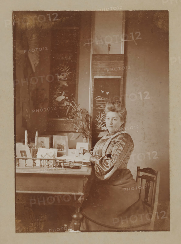 Portrait of a woman at a writing desk, photograph, Possibly a portrait of a writer., :, 1880 - 1930, photographer, paper, pasteboard, daylight collodion silver print, height 174 mm x width 122 mm, historical persons not known by name, A sepia photograph of a seated woman wearing a high-collared, long-sleeved dress with decorative trim and an elaborate updo, sitting at a wooden table with papers and small framed photos arranged on its surface, a pair of tall twisted candles visible among the items, a decorative folding screen with painted botanical and bird motifs behind her, a bouquet of foliage resting against the screen, and a wooden chair partially visible beside her.