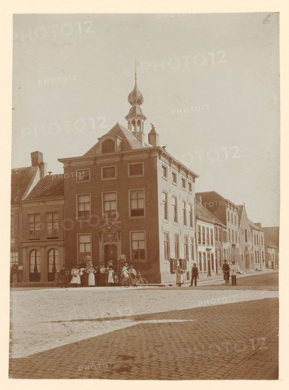 View of the town hall in Buren, photograph, annotation: 'Stadhuis, Buren', annotation: 'Het stadhuis te Buren', Buren, 1890 - 1910, photographer, paper, pasteboard, daylight collodion silver print, height 424 mm x width 295 mm, townhall, A tall brick building with a decorative entrance and multiple windows stands on a cobblestone street, topped by a roof structure with a small tower and an onion-shaped spire, flanked by neighboring buildings of varying heights and roof styles; groups of people, some standing and some seated on a bench or bicycles, gather on the sidewalk in front of the buildings, while a few individuals are seen walking or standing along the street, with shadows cast across the cobbles and an open sky above.