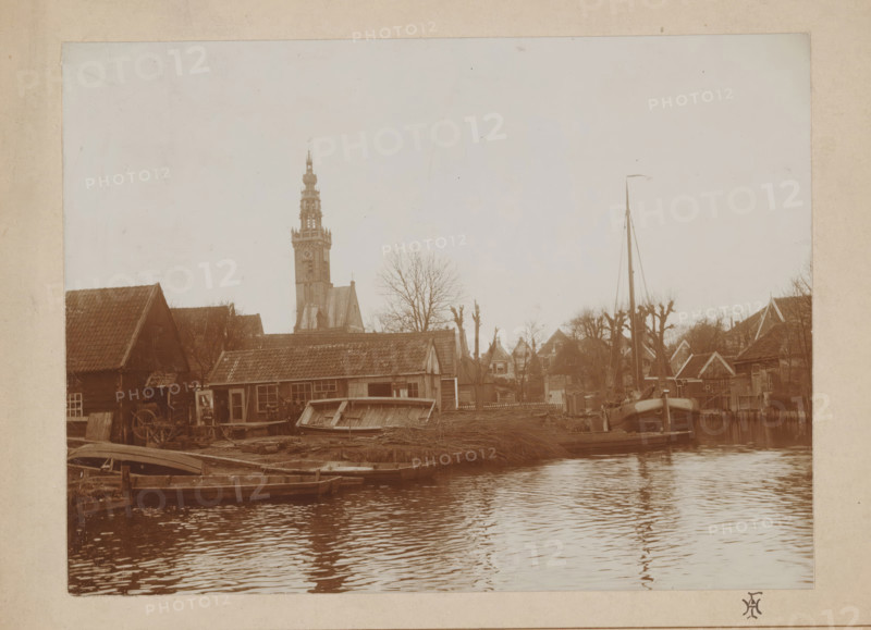 View of Edam with church tower and shipyard, photograph, monogram: 'HI', annotation: 'Scheepswerf te Edam', photographer: unknown, Edam, 1880 - 1910, paper, pasteboard, daylight collodion silver print, height 159 mm x width 208 mm, parts of church exterior and annexes: tower, village, pier, quay, wharf, A riverside scene with calm water in the foreground reflecting ripples, a wooden dock area with small boats moored along the shore, a low building with a tiled roof and a weathered facade near the water, assorted wooden structures and stacked materials on the bank, a tall church tower with ornate upper sections rising behind the buildings, leafless trees scattered among houses with pitched roofs, a tall mast with rigging and a small flag near a larger vessel, and an overall muted sepia tone across the image.