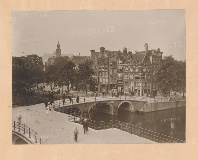 View of the Prinsengracht at the Brouwersgracht in Amsterdam, photograph, annotation: 'Prinsengracht, Brouwersgr.', annotation: 'Houses purchased by the association, Hendrik de Keyser, Prinsengracht b, d Brouwersgracht, photo ...', number: ''19.10', Amsterdam, 1900 - 1920, photographer, paper, pasteboard, gelatin silver print, developed gelatin silver print, height 244 mm x width 320 mm, urban housing, village, canals, waters (in city) (city(-scape) with figures, staffage), Prinsengracht, A sepia-toned scene shows a curved stone bridge with iron railings spanning a calm canal beneath brick arches, pedestrians walking along the cobbled street and across the bridge, blurred figures indicating motion, a row of tall narrow multi-story buildings with decorative gables and many windows lining the canal, small shopfronts at street level, leafy trees behind and between the buildings, boats moored along the canal near the trees, and a distant church tower rising above the rooftops.
