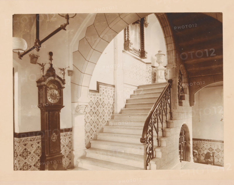 Staircase in the former Groninger Museum, photograph, annotation: 'VI, ZH, Museum Groningen', number: '1380', Groningen (city), 1880 - 1910, photographer, paper, pasteboard, daylight collodion silver print, height 299 mm x width 358 mm, staircase, A wide stone staircase with a curved wrought-iron handrail rises through an arched opening, flanked by patterned tiled walls and a stone newel supporting an ornate carved urn, while a tall grandfather clock with decorative figures and carved details stands against a tiled lower wall beneath a hanging light fixture and a ceiling beam.