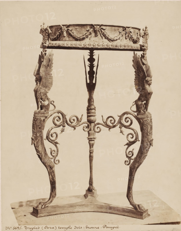 Tripod from the temple of Isis in Pompeii, Italy, Trepied (Oura) temple d'Isis: bronze: Pompei, photograph, number: '54', number: 'No. 5695', photographer: attributed to Giorgio Sommer, Pompeii, 1857 - 1914, Giorgio Sommer, photographer, Attributed to, pasteboard, paper, albumen print, height 444 mm x width 331 mm, furniture and household effects, temple, shrine, non-Christian religions, Tempio di Iside, An ornate metal stand with a circular top decorated with garland motifs rests on three elaborately carved supports; two side supports feature winged human-animal figures with detailed feathered wings and seated feline bodies atop scrollwork arms, while the central support is a vertical shaft with carved ornamentation and a stylized silhouette at its upper section; the lower ends of the supports terminate in curved legs with pawed feet standing on a rectangular base, and the piece is shown against a plain light background.