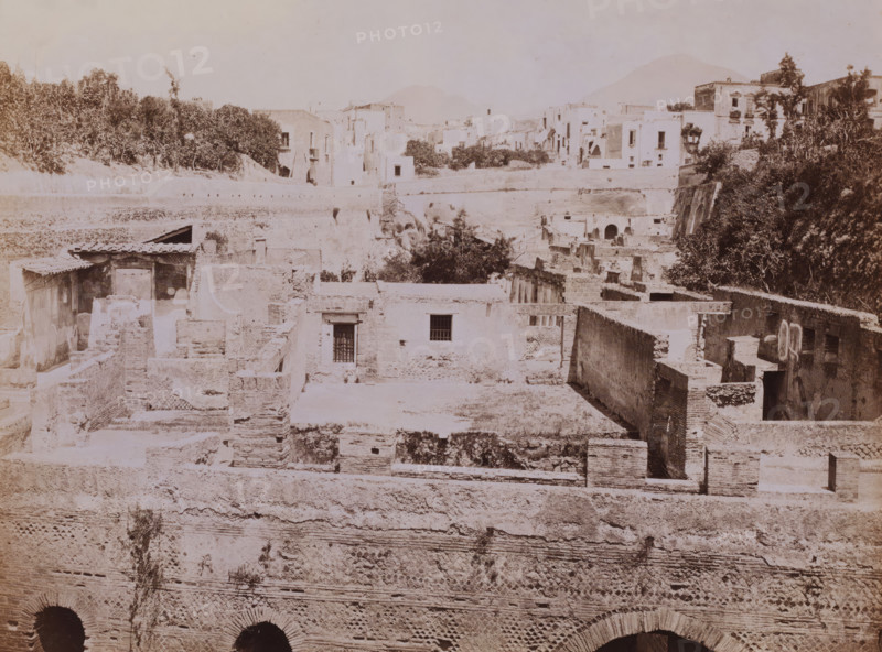 Panorama di Ercolano, View of ruins at Herculaneum, Italy, photograph, number: 'No. 1291', collector's mark: 'VOSMAER', photographer: Giorgio Sommer, Herculaneum, 1857 - 1875, Giorgio Sommer, photographer, pasteboard, paper, albumen print, height 315 mm x width 421 mm, urban housing, city-view in general, 'veduta', ruin of a building, architecture, destroyed, ruined place or city (, warfare), A sepia-toned scene of layered stone ruins and buildings with flat roofs and rectangular openings, showing exposed brickwork, archways, and remnants of walls at various heights; trees and dense foliage appear on the right and along the upper terrace, with more clustered buildings and terraces in the background; the foreground features textured masonry with patterned brick sections and dark arched openings at the bottom, while scattered stairways, small barred windows, and a few roofed shelter structures are visible among the crumbling platforms and courtyards.
