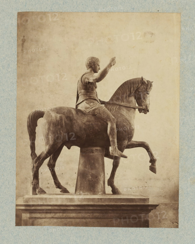 Equestrian statue, possibly of a Roman emperor, photograph, collector's mark: 'VOSMAER', Italy, after sculpture by anonymous, 1851 - 1890, photographer, after sculpture by, pasteboard, paper, albumen print, height 325 mm x width 249 mm, equestrian statue, A sepia-toned photograph of an equestrian statue showing a figure seated on a horse, the rider wearing a short tunic and cuirass with one arm raised and the other holding the reins, the horse depicted with a lifted foreleg and detailed mane and tail, the sculpture mounted on a cylindrical support above a rectangular pedestal, the image framed by a textured light-colored border.