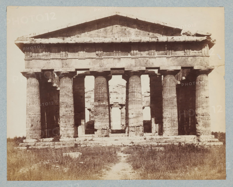 Tempio di Nettuno. Pesto, Frontal view of the Temple of Neptune in Paestum, Italy, photograph, collector's mark: 'VOSMAER', inscription, photographer: attributed to Giorgio Sommer, Paestum, 1857 - 1914, Giorgio Sommer, photographer, Attributed to, pasteboard, paper, albumen print, albumendruk, height 250 mm x width 325 mm, ruin of a building, architecture, temple, shrine, non-Christian religions, Second Temple of Hera, An aged stone structure with a triangular pediment atop a horizontal entablature sits on a raised stepped platform, supported by a row of fluted columns with simple capitals, some columns showing weathered and eroded surfaces, and gaps between them revealing more ruins behind; the foreground shows uneven grassy ground and scattered stone blocks leading to the structure, and the whole scene is framed by a pale border.