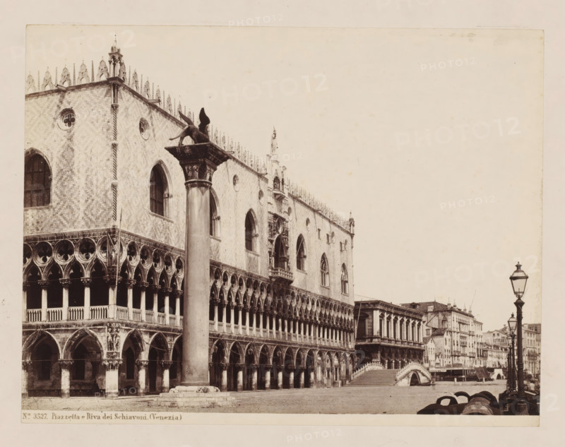 View of the Piazzetta and Riva dei Schiavoni in Venice, Italy, Piazzetta e Riva dei Schiavoni. (Venezia), photograph, inscription: 'Box 22, = Sommer (van 1182-W)', inscription: 'Venice Doge Palace', number: 'No. 3527.', inscription: 'I', collector's mark: 'RIJKS SCHOOL FOR ART NIJVERINGEN, GROEP', inscription: 'H', photographer: attributed to Giorgio Sommer, Venice, 1857 - 1914, Giorgio Sommer, photographer, Attributed to, pasteboard, paper, albumen print, albumendruk, height 319 mm x width 479 mm, square, circus, Piazzetta, Riva degli Schiavoni, A sepia-toned photograph of an ornate Gothic building with arched colonnades and patterned masonry, showing multiple rows of pointed arches, circular windows, and decorative stone tracery, a tall column topped by a winged lion sculpture standing in front of the facade, a small bridge leading to another building with classical columns, a row of street lamps and waterfront buildings receding into the distance, barrels and mooring posts along the...