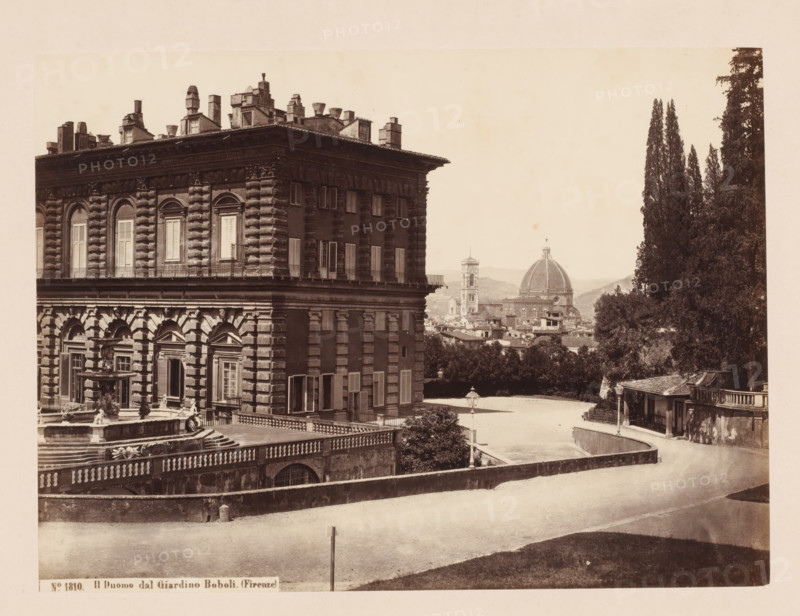 Il Duomo dal Giardino Boboli. (Firenze), View of the Pitti Palace, the Boboli Gardens and the Duomo of Florence, Italy, photograph, address: 'SOMMER E BEHLES, NAPOLI Monte di Dio 4, DEPOT A FLORENCE, VIA MAGGIO 15', number: 'No. 1810.', photographer: Sommer & Behles, Florence, 1867 - 1874, Tuscany, pasteboard, paper, albumen print, height 307 mm x width 375 mm, park, church (exterior), tower (of house or building), hôtel, ie small palace in city, Florence Cathedral, Palazzo Pitti, Giardino di Boboli, A sepia-toned scene showing the corner of a large ornate stone building with rusticated masonry, tall shuttered windows, decorative cornices, and a balustraded terrace with a sculpted fountain in front; a broad paved area and curved driveway run alongside the building, bordered by low stone walls and a lamppost, with a cluster of tall cypress trees and other foliage rising on the right; in the middle distance a domed cathedral and a bell tower rise above a cityscape of tiled roofs and dist...