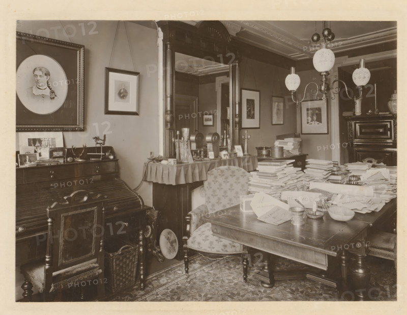 Study of Pieter Lodewijk Tak, photograph, photographer: Friedrich Carel Hisgen, Amsterdam, 1884 - 1907, paper, pasteboard, baryta paper, gelatin silver print, developed gelatin silver print, height 295 mm x width 361 mm, Person, Pieter Lodewijk Tak, interior of the house, A sepia-toned interior room filled with antique furniture and many stacks of papers on a large wooden table, showing ornate wooden chairs with patterned upholstery, a carved desk with a roll-top surface, and a wicker basket on the floor; framed portraits and pictures hang on the walls, including a large oval portrait of a woman, and smaller rectangular frames suspended by cords; a tall wooden mirror with decorative columns stands behind a side table covered with a draped cloth holding candlesticks, small framed photos, and decorative objects; a multi-arm ceiling lamp with frosted glass globes hangs above the table, and a patterned rug covers the floor beneath the furniture.