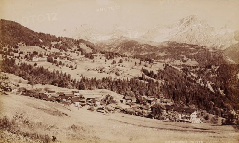 Chesieres Villars, View of Villars-sur-Ollon in Switzerland, photograph, number: '1781', collector's mark: 'VOSMAER', Villars-sur-Ollon, 1900, photographer, paper, albumen print, albumendruk, height 132 mm x width 221 mm, city view in general, 'veduta', mountains, A wide sepia-toned landscape showing a mountain valley with clustered buildings and houses in the foreground along a sloping grassy hillside, a band of dense conifer forest rising behind them, scattered individual trees and small clearings across rolling meadows in the middle ground, larger multi-story buildings on a distant slope, and rugged snow-dusted mountain peaks and ridgelines forming the background under a pale sky.