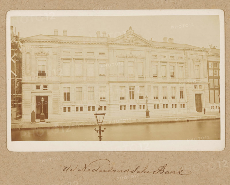 Façade of Oude Turfmarkt 127 in Amsterdam, former De Nederlandsche Bank, photograph, annotation: 'Nederlandsche Bank', Amsterdam, 1870 - 1890, photographer, paper, pasteboard, albumen print, height 94 mm x width 157 mm, height 162 mm x width 251 mm, bank, civic architecture, edifices, dwellings, Rokin, Oude Turfmarkt, A sepia-toned photograph of a large classical stone building with tall windows and decorative pediments along its facade, a triangular sculpted pediment above the central section, and chimneys visible on the roof; a canal runs in front of the building with a decorative street lamp reflected in the water, a second lamp visible on the sidewalk beside the building, scaffolding is present at the far left edge, and a solitary figure stands near a large doorway on the right side.