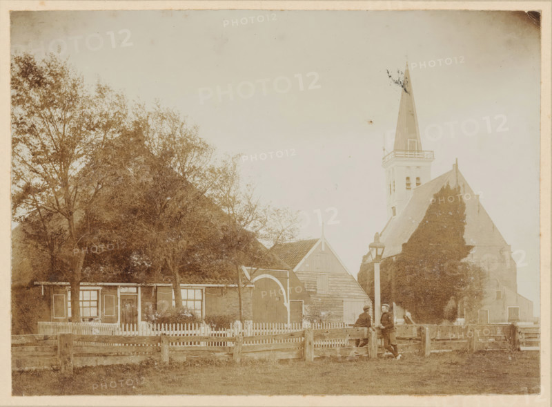 View of a farm and the Reformed Church in Den Hoorn, Texel, photograph, annotation: 'Church in Den Hoorn Texel', annotation: '16 1, 2', Den Hoorn (Texel), 1890 - 1910, photographer, paper, pasteboard, daylight collodion silver print, height 119 mm x width 163 mm, height 219 mm x width 269 mm, church (exterior), farm (building), A faded sepia photograph shows a village scene with a row of buildings partially obscured by tall leafy trees on the left and a steep church roof and tall pointed steeple on the right. In front of the buildings a picket fence runs horizontally, interrupted by a wooden gate and a larger arched doorway set into a barnlike structure. A lamp post stands near the center right beside a low wooden fence and two men wearing caps and coats lean against the fence facing each other. The ground in the foreground is grassy and the sky above is pale and empty.