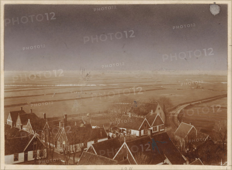 View of Oosterend in Texel, photograph, annotation: '13 1, 2', annotation: 'Oosterend, Texel', Oosterend, 1890 - 1910, photographer, Texel, paper, pasteboard, daylight collodion silver print, lithography, daylight collodion silver print and lithography, height 120 mm x width 165 mm, height 220 mm x width 270 mm, agriculture, village, A sepia-toned aerial view shows a cluster of pitched-roof houses and buildings in the foreground with visible chimneys, windows, and roof tiles, some trees without leaves scattered among them, fences and narrow pathways between the structures, and laundry or cloth hanging on a line near one house; beyond the houses stretches a flat, open landscape of fields and waterways with linear dikes and meandering roads, a few small isolated structures in the distance, and a wide sky above that fades from darker tone at the top to lighter near the horizon.