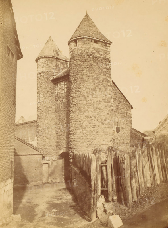 View of the Helpoort in Maastricht, photograph, inscription: 'Helpoort Maastricht', name: 'S. FRANCO, bi The Hague.', photographer: Héliodore Dandoy, Maastricht, c. 1875 - in or before 1884, paper, pasteboard, albumen print, height 222 mm x width 165 mm, city-gate, façade (of house or building), gate, entrance, Helpoort, A sepia-toned photograph of a stone building with two conical-roofed cylindrical towers, rough stone walls, and small window openings, flanked by brick structures on the left and a wooden palisade fence on the right, with a narrow dirt pathway leading toward an arched entrance at the base of the central tower and shadowed areas along the ground.
