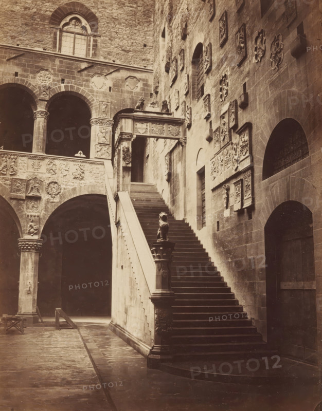 Stairs and courtyard of the Palazzo del Bargello in Florence, photograph, Florence, c. 1875 - c. 1900, photographer, Tuscany, paper, pasteboard, albumen print, albumendruk, height 250 mm x width 195 mm, courtyard, palace, staircase, A sepia photograph of a stone courtyard featuring a wide staircase with a solid balustrade leading upward to an ornate covered walkway, a sculpted lion figure atop a newel at the base of the stairs, and a series of arched openings and columns with carved capitals along the left side; the surrounding walls display numerous decorative relief panels and sculpted medallions, with a large arched doorway visible on the right and a tall window with a rounded top set high on the wall above the arches.