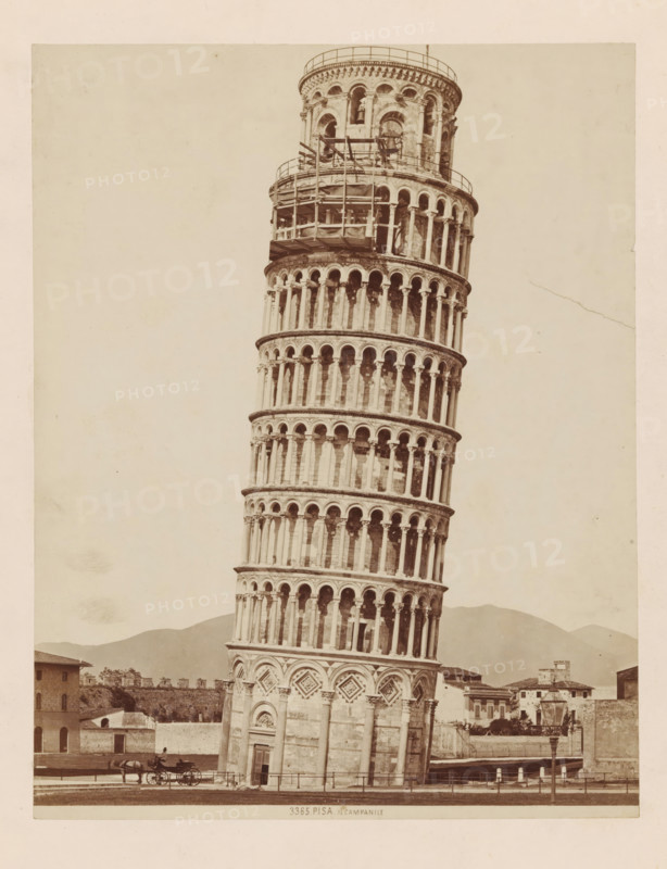 Tower of Pisa, PISA . Il Campanile, photograph, annotation, number: '3365.', Pisa, c. 1875 - c. 1900, photographer, Tuscany, paper, pasteboard, albumen print, albumendruk, height 247 mm x width 193 mm, four-wheeled, animal-drawn vehicle, eg: cab, carriage, coach, tower, clock tower, monumental adornment, A tall, leaning stone tower with multiple tiers of arched openings and decorative columns rises from a base featuring ornate geometric panels and a doorway; scaffolding and a railing are visible near the upper portion of the tower. The foreground shows a paved area with a horse-drawn carriage and a carriage driver near the tower base, along with a low fence and a lamppost. Surrounding the tower are low buildings and walls, and distant hills form the backdrop beneath a pale, empty sky.