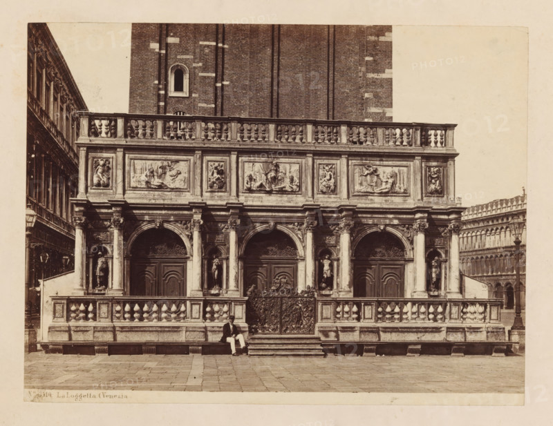 Loggetta del Sansovino in Venice, La Loggetta (Venezia, photograph, number: 'No. 3910.', Venice, c. 1875 - c. 1900, photographer, paper, pasteboard, albumen print, albumendruk, height 180 mm x width 237 mm, column, pillar, architecture, facade (of house or building), A sepia-toned photograph of an ornate stone building facade with a balustraded upper terrace and a series of carved rectangular panels depicting sculptural reliefs. The lower level shows three large arched doorways with decorative metalwork gates and flanking niches containing full-length statues. A sculpted railing runs along the terrace above a broad flight of shallow steps. A seated man in dark jacket and light trousers is positioned on the steps near the center. Flanking wings of colonnaded architecture extend to the left and right edges of the image, and a tall brick wall with a single arched window rises behind the balustrade. The foreground is a paved stone plaza.