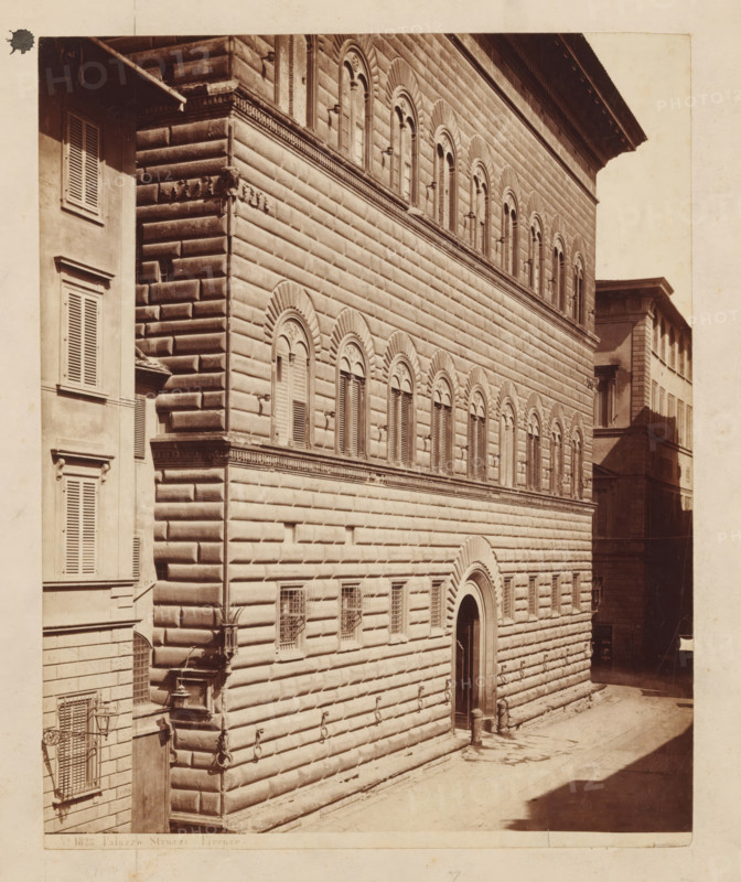 Façade of Palazzo Strozzi in Florence, Palazzo Strozzi Firenze, photograph, number: 'No. 1823.', Florence, c. 1875 - c. 1900, photographer, Tuscany, paper, pasteboard, albumen print, height 247 mm x width 197 mm, palace, façade (of house or building), A sepia-toned photograph showing the facade of a large stone building with rusticated masonry and rows of arched windows with decorative surrounds, a tall arched doorway set into the wall, smaller barred windows near the ground, and metal rings attached to the lower stone courses; adjacent on the left is a narrower building with shuttered windows and a small awning, while a shadowed narrow street runs alongside the right side and another building with shuttered windows is visible in the background.
