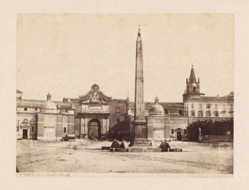 View of Porta del Popolo and the Flaminische Obelisk in Rome, Porta del Popolo (Roma), photograph, number: 'No. 1009.', Rome, c. 1875 - c. 1900, photographer, paper, pasteboard, albumen print, height 178 mm x width 237 mm, square, circus, city-gate, obelisk, needle, A wide sepia photograph of a plaza featuring a tall, narrow obelisk with carved markings standing on a stepped stone base and topped with a small cross, flanked by classical buildings with domed roofs and decorative facades, an arched gateway with sculptural ornament above it, a rectangular building with a clock tower and spire to the right, and a small statue on a pedestal near a group of trees, all set on a broad open paved foreground.