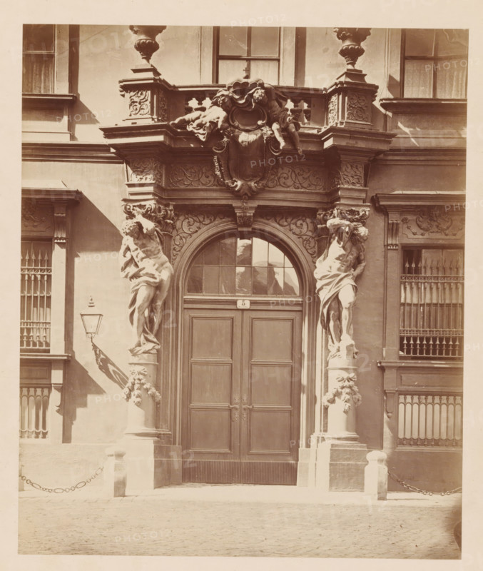 Fürstllich Liechtenstein'sches Palais, SEITEN-PORTAL', Side entrance of Stadtpalais Liechtenstein in Vienna, photograph, number: 'Nr. 48.', Vienna, publisher: Ludwig Niernberger, c. 1875 - c. 1900, photographer, Lower Austria, paper, pasteboard, albumen print, height 235 mm x width 197 mm, piece of sculpture, reproduction of a piece of sculpture, coat of arms (as symbol of the state, etc.), gate, entrance, A sepia-toned photograph of a building entrance with a large double wooden door set beneath an arched transom window, flanked by two tall sculpted male figures carved as supporting columns, ornate capitals, and decorative floral garlands on the column bases; above the arch is an elaborate carved entablature with scrollwork and a central cartouche topped by reclining cherub-like figures and decorative urns on pedestals; to either side of the entrance are windows with vertical bars and decorative moldings, a wall-mounted lantern with a bracket casting a shadow, short stone posts connec...