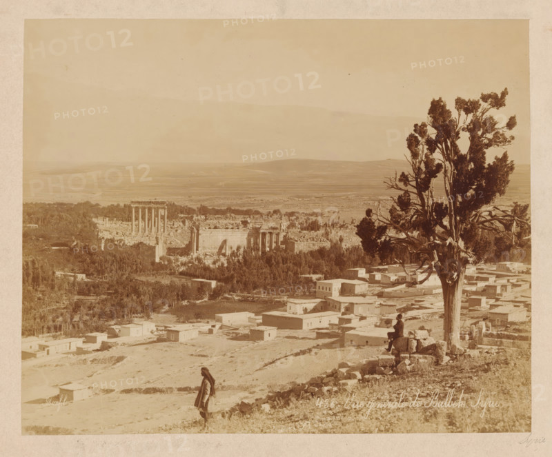View of Baalbek, Vue générale de Balbek. Syrie., photograph, name: 'JAG DE LEUR Représentant Huigenspark, 40b. THE HAGUE.', number: '438', photographer: Maison Bonfils, Baalbek, c. 1875 - c. 1900, paper, pasteboard, albumen print, height 212 mm x width 262 mm, trees, landscape with ruins, A sepia-toned panoramic view showing an expansive landscape with ruins of a large columned temple complex near the center left, scattered rectangular buildings in the foreground, and a cluster of trees and vegetation around the ruins. A tall, gnarled tree with dense foliage stands on the right side, its trunk and branches prominent against the sky. A person wearing traditional clothing walks along a slope in the lower foreground, and another person sits on a low stone wall near the base of the tree. Rolling plains and distant low hills extend across the background beneath a pale sky.