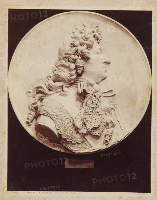 P. Puget Louis XIV. Musée de Marseille., Relief with profile portrait of King Louis XIV, by P. Puget, photograph, number: 'T 427', collector's mark: 'RIJKS-SCHOOL VOOR TEEKENONDERWIJZERS AMSTERDAM', photographer: Adolphe Giraudon, c. 1875 - c. 1900, Adolphe Giraudon, photographer, paper, pasteboard, albumen print, height 260 mm x width 204 mm, sculpture, historical persons ((full) bust portrait), sculpture (relief, sculpture), A circular relief portrait of a man in profile facing right set against a dark rectangular background, showing detailed curly hair cascading past his shoulder, a prominent nose and chin, and an ornate lace cravat and decorative armor with carved patterns and flourishes, all rendered in a monochrome stone or plaster tone with small specks and slight discolorations on the surface.