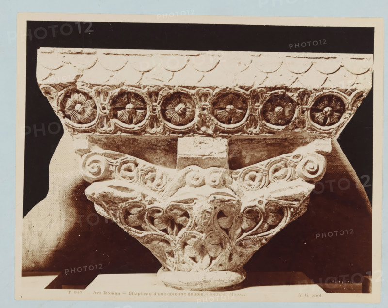 Capital of a double column with plant motifs, Art Roman - Chapiteau d'une colonne double. Cloître de Moissac., photograph, number: 'T 947', collector's mark: 'INSTITUUT VOOR KUNST-NIJVERHEIDSONDERWIJS GABRIËL METSUSTRAAT No. 16, AMSTERDAM', collector's mark: 'Dagteeken- en Kunstambacht- school voor Meisjes Amsterdam', name: 'A. GIRAUDON ÉDITEUR 15. R. BONAPARTE. PARIS', photographer: Adolphe Giraudon, publisher: Adolphe Giraudon, Paris, c. 1875 - c. 1900, Adolphe Giraudon, photographer, publisher, paper, pasteboard, albumen print, height 205 mm x width 260 mm, flowers, ornament, foliage, tendrils, branches, capital (, column, pillar), sculpture (relief, sculpture), A close-up photograph of an ornate carved stone capital showing a row of circular floral motifs along the top band, each set within a carved roundel, beneath a plain molded abacus with scalloped detailing; below the band a central rectangular block sits above a triangular arrangement of carved foliage and spiral volutes with...