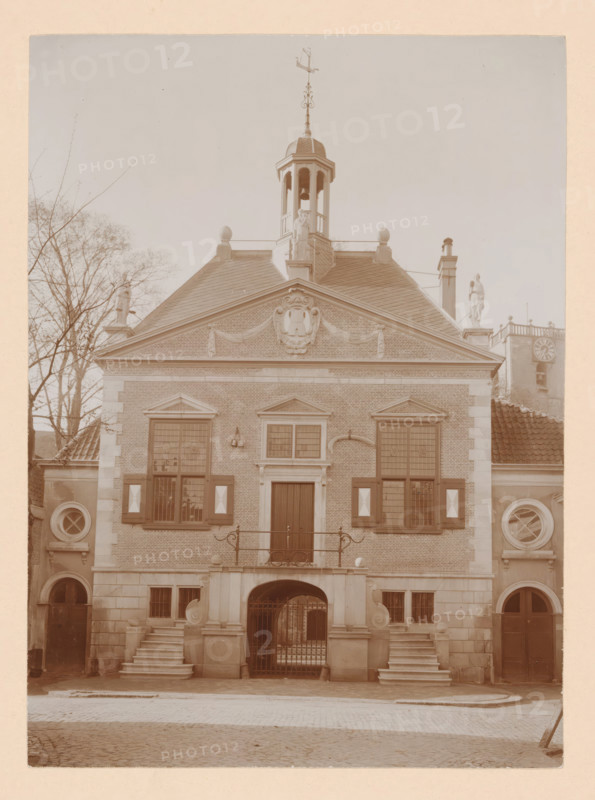 Middelharnis Town Hall, photograph, annotation: 'Town Hall, after the restoration 1909, Middelharnis', annotation: 'C. van Rhijn, 1683-1745', Middelharnis, 1909, photographer, Attributed to, paper, pasteboard, daylight collodion silver print, height 225 mm x width 163 mm, town hall, façade (of house or building), A symmetrical brick building facade with a triangular pediment topped by a small cupola and a decorative weather vane, flanked by statues on the roof corners and a tree visible to one side; shuttered windows with patterned panels and circular windows set into stone sections at either side; a central doorway set above a short staircase and balcony with wrought iron railing, an arched gated passageway beneath, and paired stone staircases leading to raised entrances; cobblestone street in front and a tall chimney and clock tower partially visible beyond one roofline.