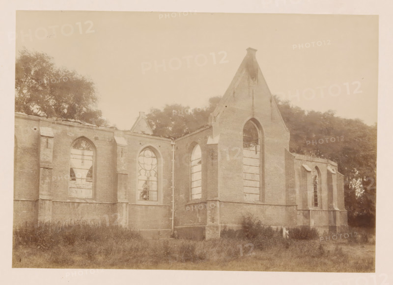 View of the Reformed Church in Middelharnis after the fire, photograph, annotation: 'Middelharnis (ZH), Kerk der Ned. Rev. congregation, on July 5, 1904', annotation: 'Kerk der Ned. Rev. Municipality, on 5 July 1904, Middelharnis', Middelharnis, 1904, photographer, Attributed to, paper, pasteboard, albumen print, albumendruk, height 164 mm x width 230 mm, church (exterior), A sepia-toned photograph showing the exterior of a large brick building with tall arched window openings, many of which are missing glass and have visible wooden supports; the building has buttresses and a steep gable end with a narrow top, and a vertical drainpipe runs down one corner; trees and foliage appear behind and to the sides of the structure, and unkempt grass fills the foreground.