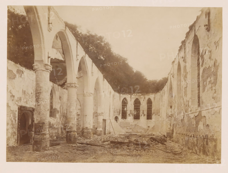 Interior of the Reformed Church in Middelharnis after the fire, photograph, annotation: 'Middelharnis (ZH), Church of the Dutch Reformed congregation on July 5, 1904, viewed towards the east', annotation: 'Church of the Dutch Reformed Congregation on July 5, viewed towards the East, Middelharnis', Middelharnis, photographer, Attributed to, paper, pasteboard, albumen print, height 163 mm x width 225 mm, interior of church, A large interior space with tall, arched openings and rows of columns showing worn and chipped surfaces, plaster peeling from the walls, and rubble scattered across the floor; high windows with pointed tops are set into a curved far wall, and remnants of beams and metal rods lie among debris; the ceiling is absent, revealing treetops and sky above the upper edges of the walls, and the overall scene shows faded, uneven surfaces and architectural detailing around the arches.