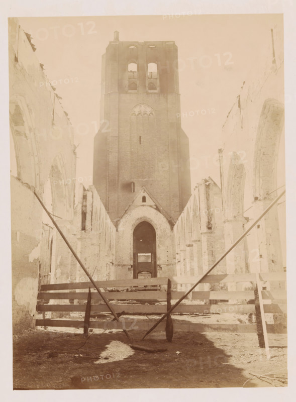 Interior of the Reformed Church in Monster after the fire, photograph, annotation: 'Monster NH Church (ZH), view to the west after the fire July 1901', annotation: 'NH Church, viewed to the West, after the fire, July 1901, Monster', Monster (South Holland), 1901, photographer, Attributed to, paper, pasteboard, albumen print, height 230 mm x width 170 mm, interior of church, A tall brick tower with large arched openings stands at the center of the image, flanked by the ruined walls of a long building with rows of pointed arch openings on both sides; the interior foreground shows wooden beams and planks laid across the space with angled support poles crossing toward the viewer, rubble and uneven ground visible beneath, and light filtering through the empty archways toward the distant tower.