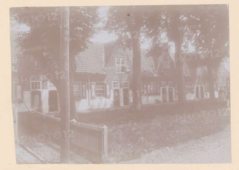 Courtyard of the Old Men's House in Naaldwijk, photograph, annotation: 'Facades of the Old Men's House to the west of the courtyard, May 1910, Naaldwijk', Naaldwijk, 1910, photographer, Attributed to, paper, pasteboard, daylight collodion silver print, height 157 mm x width 219 mm, courtyard, façade (of house or building), A row of brick houses with tiled gabled roofs is partially obscured by tall trees in front; each house has shuttered windows and small doorways, and some windows have visible windowboxes. A wooden picket fence runs along a grassy area in front of the houses, and a cobblestone path or street is visible in the foreground. A wooden pole stands near the fence, and tree trunks and leafy canopies cast shadows across the scene. A few figures are faintly visible near the doorways along the row of houses.