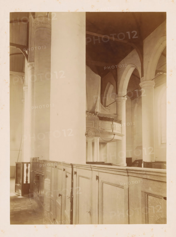 Interior of the Reformed Church of Noordwijk-Binnen, photograph, annotation: 'NH Church, interior, July 1901, Noordwijk-Binnen', annotation: 'Noordwijk-binnen (ZH), NH church, July 1901', Noordwijk-Binnen, 1901, photographer, Noordwijk, Attributed to, paper, pasteboard, albumen print, height 230 mm x width 169 mm, interior of church, A sepia-toned interior view showing tall rounded columns and arches, a high ceiling with visible beams, a wooden balcony with ornate railing partly draped in fabric, a paneled wooden partition or pews running along the foreground with small doors and metal fixtures, a section of a stair or railing at the left, stone or tiled flooring, and soft diffused light coming through tall arched windows.