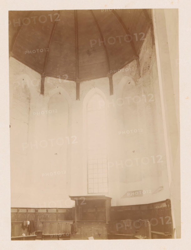 Choir of the Reformed Church of Noordwijk-Binnen, photograph, annotation: 'Noordwijk-binnen (ZH), NH church, choir view, July 1901', annotation: 'NH Church, July 1901, Noordwijk-Binnen', Noordwijk-Binnen, 1901, photographer, Noordwijk, Attributed to, paper, pasteboard, albumen print, height 230 mm x width 169 mm, interior of church, A high interior space with a domed ceiling supported by visible ribs above tall arched wall openings, pale walls showing patches of wear, a tall narrow window with a grid of panes letting diffuse light into the space, wooden paneling and benches along the lower walls, a raised wooden pulpit or lectern at the center front, and various cloths draped over the benches and pulpit.