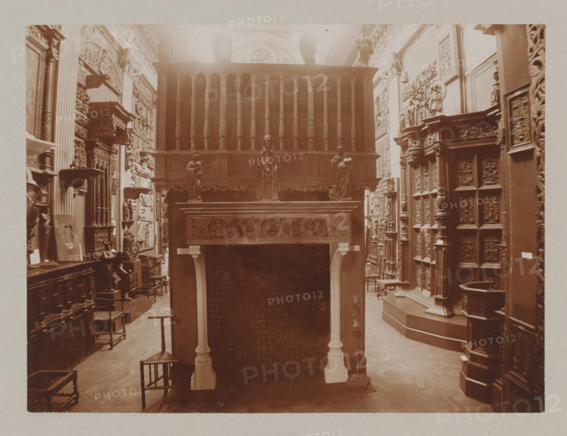 View of a hall with a fireplace and reliefs, album page, photograph, This album page is part of a cover with 6 pages., collector's mark: 'Museum van Kunst-Nijverheid Haarlem', workshop stamp: 'J. Pörtzgen, Photograaf, Koningstraat 38, Haarlem', photographer: Johann Pörtzgen, c. 1890 - c. 1900, Johann Pörtzgen, photographer, paper, pasteboard, daylight collodion silver print, height 166 mm x width 221 mm, open hearth, fire-place, ornamenting, decorating, applied arts, A sepia-toned interior scene showing a room filled with carved wooden furniture and decorative panels, featuring a tall ornate wooden structure with turned balusters and a carved frieze, three small statues positioned on a raised ledge, an elaborately carved mantel with floral and circular motifs supported by slender columns, rows of intricately detailed panels and pilasters lining the walls, several small chairs and stools on a wooden floor, sculpted reliefs and framed carved plaques mounted on the walls, and various orna...