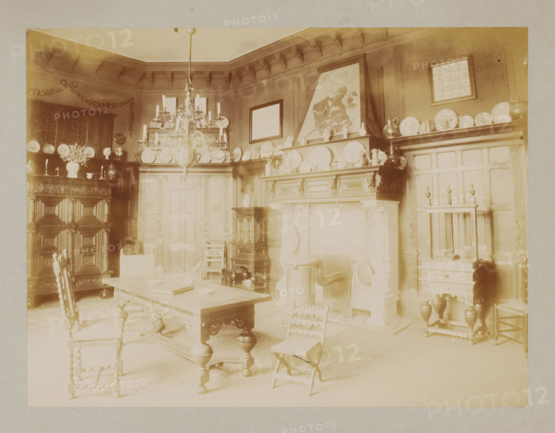 Interior with a desk, chairs and decorative plates, album page, photograph, This album page is part of a cover with 6 pages., workshop stamp: 'J. Pörtzgen, Photographer, Koningstraat 38, Haarlem', collector's mark: 'Museum van Kunst-Nijverheid Haarlem', photographer: Johann Pörtzgen, c. 1890 - c. 1900, Johann Pörtzgen, photographer, paper, pasteboard, daylight collodion silver print, height 166 mm x width 220 mm, chandelier, candelabrum, interior of the house, chair, A sepia-toned interior room with ornate wood paneling and carved furniture, featuring a large wooden table with turned legs at the center and chairs with twisted spindle backs around it, a chandelier hanging from the ceiling above the table, shelves and mantels lined with decorative plates and pitchers, a prominent carved fireplace surround with decorative reliefs and objects on its mantel, framed artworks leaning and hanging on the walls above the mantel, a tall cabinet with intricately carved doors, smaller cabinets and ...